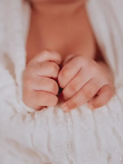 A close-up of a newborn's tiny fists, nestled together. The soft texture of the white swaddle and the orange blanket create a cozy and warm feeling.