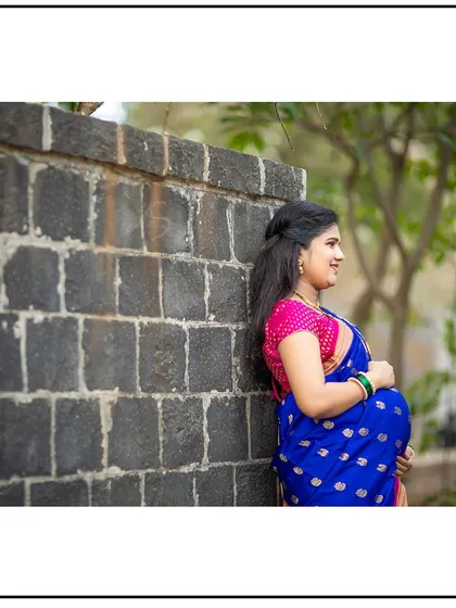 A contemplative side-profile of a mom-to-be in her beautiful blue saree, leaning against a stone wall.