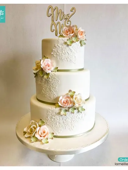 This classic three-tiered wedding cake exudes timeless elegance. It features intricate white-on-white lace piping, subtle ribbon bands, and is topped with a custom 'Mr & Mrs' topper and delicate sugar roses.
