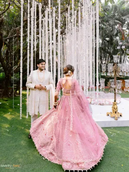 The bride twirls in her beautiful pink lehenga as she approaches her groom, who waits for her amidst a curtain of white flowers. A moment of joyful anticipation.
