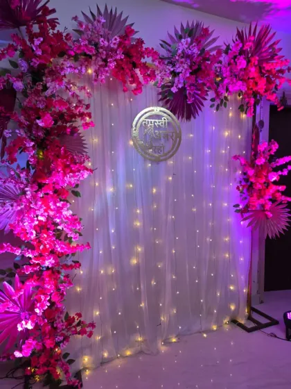 A vibrant pink floral arch against a white curtain backdrop with fairy lights. The golden "Tapasvi Amar Raho" sign adds a touch of elegance to this home ceremony setup.