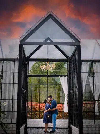 A romantic kiss in the doorway of a modern glasshouse under a fiery sunset sky. A stunning and epic pre-wedding photograph.