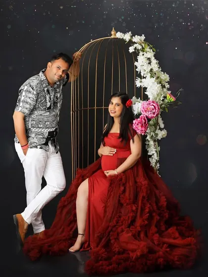 This artistic portrait features a golden birdcage prop adorned with flowers. The couple poses elegantly, with the mom-to-be seated in a dramatic red gown, creating a fairytale-like scene.