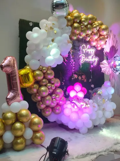 A fun and stylish first birthday party decoration. The setup includes a circular balloon arrangement in gold and white, a 'Happy Birthday' neon sign, and a large '1' balloon number.