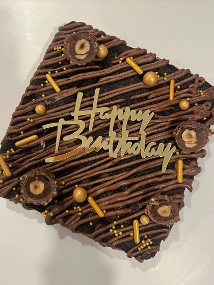A close-up of my Ferrero Rocher brownie cake, perfect for a birthday celebration. It's drizzled with chocolate and topped with crunchy hazelnuts, sprinkles, and a "Happy Birthday" plaque.