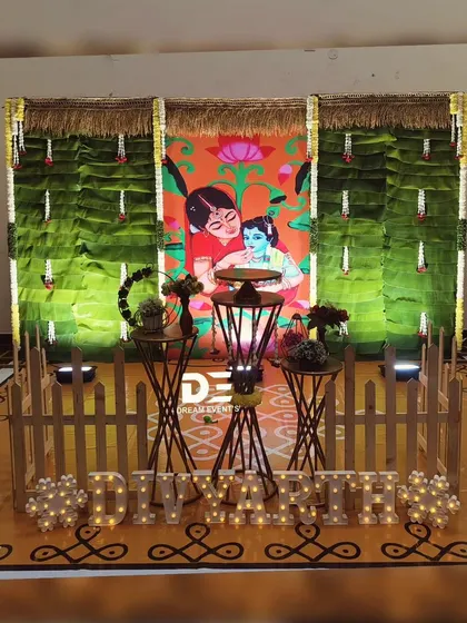 A unique naming ceremony setup featuring a custom printed backdrop of Yashoda and Krishna, framed by banana leaves and marquee lights spelling out the baby's name.