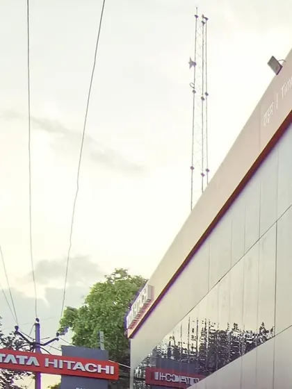The exterior facade of the Tata Hitachi building, showing the clean lines of the charcoal and silver aluminum composite panels. The prominent red branding element creates a strong visual landmark.