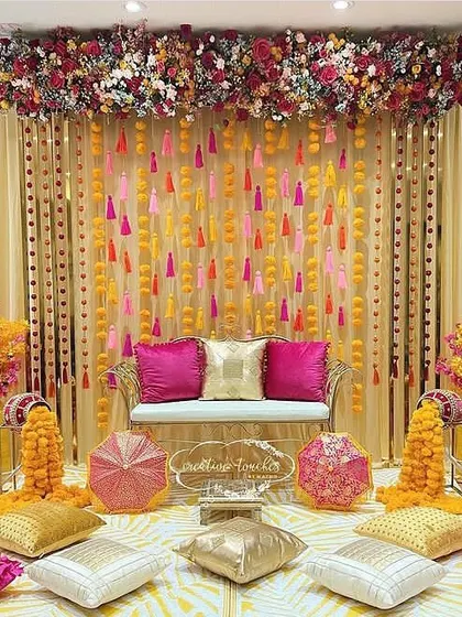 A luxurious indoor Mehendi setup. The decor features a rich color palette of gold and pink, with hanging tassels, marigolds, and plush seating.