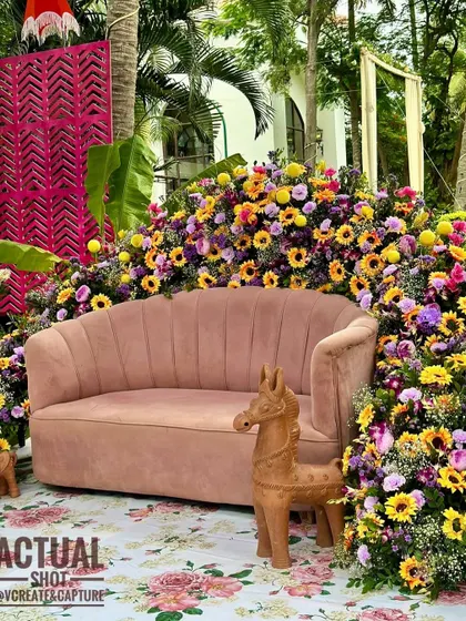 A beautiful seating area for a Haldi or Mehendi, with a plush pink sofa surrounded by a sea of sunflowers and other colorful blooms.