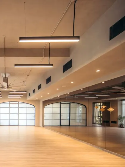 An expansive view of the yoga studio, highlighting the high ceilings and industrial-style fans. The large, arched windows with black frames add a touch of classic elegance, while the full-length mirror wall visually doubles the space.