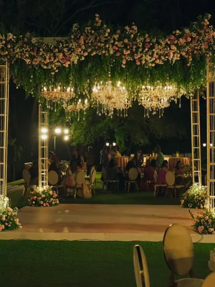 As day turns to night, the floral canopy comes alive with the warm glow of chandeliers, creating an enchanting atmosphere for an evening of celebration.