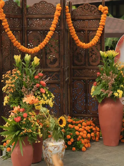 A cozy corner at a Haldi event, featuring a traditional carved wooden screen, marigold garlands, and earthy terracotta pots filled with colorful flowers.