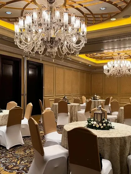 A view of the banquet hall at The Leela Palace, Delhi, set up for a luxury event. The room is filled with elegantly set tables under magnificent crystal chandeliers.