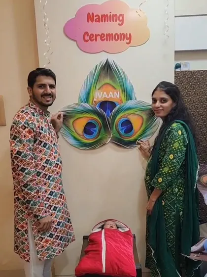 A happy family posing with our custom decor for their baby's naming ceremony. We created a personalized wall hanging with a 'Naming Ceremony' sign and peacock feather artwork incorporating the baby's name, 'Ivaan'.