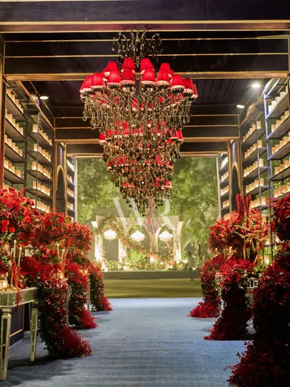 The grand entrance to the red and blue themed event. A long corridor lined with overflowing red florals and candle walls, under a massive red chandelier, creates an unforgettable first impression.