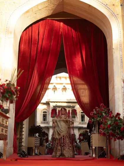 A dramatic bridal entry through heavy red velvet curtains, creating a sense of royal theatre and anticipation. The architecture of the palace is used to frame this unforgettable moment.