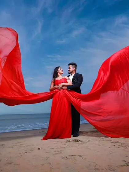 A duplicate of image 19, showing the full beach scene. The groom's classic black suit provides a sharp, elegant contrast to the flowing red gown and the blue sky.