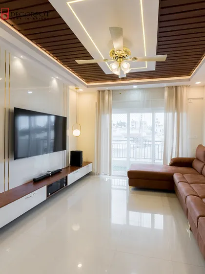 This bright and airy living room features a TV unit with a white back panel and gold accents, set against a wooden false ceiling. The design maximizes natural light from the large balcony doors.