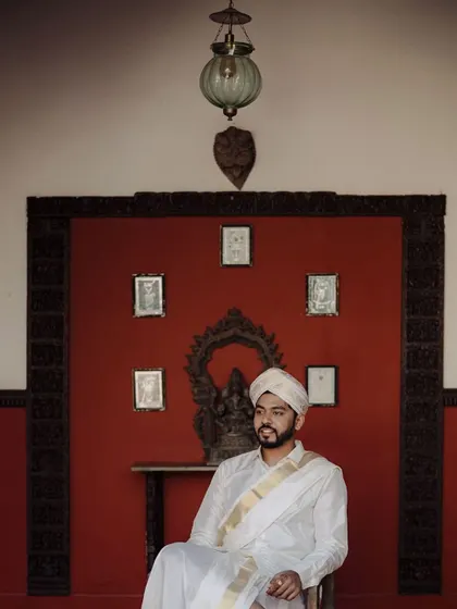 An elegant portrait of the groom in traditional Kannadiga attire. The setting, with its classic red wall and traditional decor, adds a sense of heritage and dignity to the shot.