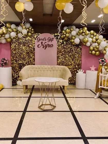 A glamorous baptism decor setup in pink, white, and gold. The stage features a gold shimmer backdrop, balloon arches, and a decorated cradle for the baby.