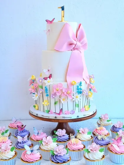 A beautiful two-tier first birthday cake with a garden theme, complete with a large pink fondant bow. This set includes matching floral cupcakes, showing how I can create a cohesive and elegant dessert table for your celebration.
