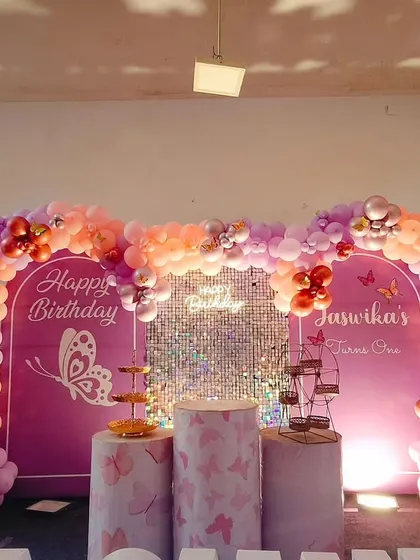 A butterfly theme birthday setup with a double-arch backdrop in purple. The balloon arrangement in shades of purple, peach, and red creates a warm and inviting look, complemented by the central sequin panel.