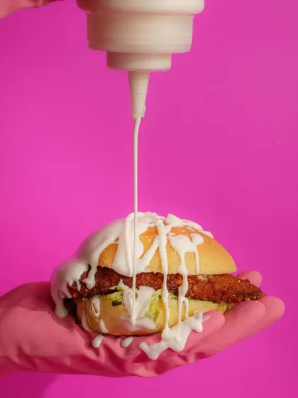 A satisfying sauce drizzle on a burger, but with a twist. The pink glove adds a pop of color and a touch of surrealism, elevating a simple action into a piece of pop art.