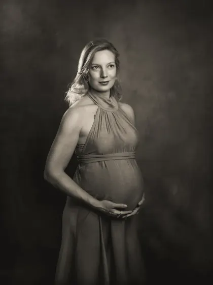 A duplicate of ID 19, this is a contemplative pose from a studio maternity session. The expecting mother looks down at her belly, her hands cradling it gently, against a painterly, textured backdrop. This is the B&W version.