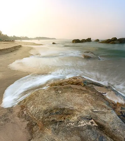 A long exposure captures the motion of waves washing over rocks on a sandy beach. The glistening sunlight dances on the rolling waves, creating the ocean's endless song.