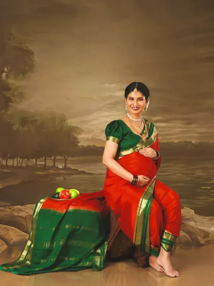 A divine maternity portrait with a painted background, giving it a classic, artistic feel. The mom-to-be looks radiant in her traditional red and green saree, embodying grace and beauty.