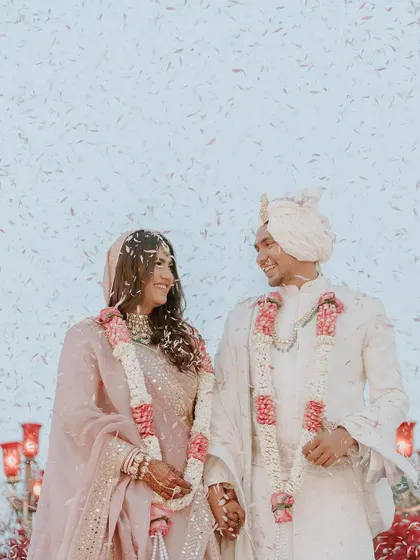 The couple shares a smile under a shower of white petals, with the vibrant red floral decor creating a striking and romantic frame.