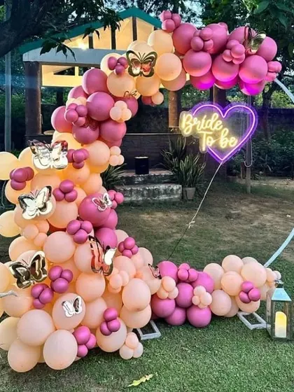 Perfect for a garden or lawn party, this crescent-shaped balloon arch uses a mix of pastel and bright pink balloons. The setup is completed with a heart-shaped 'Bride to be' neon sign and decorative lanterns.