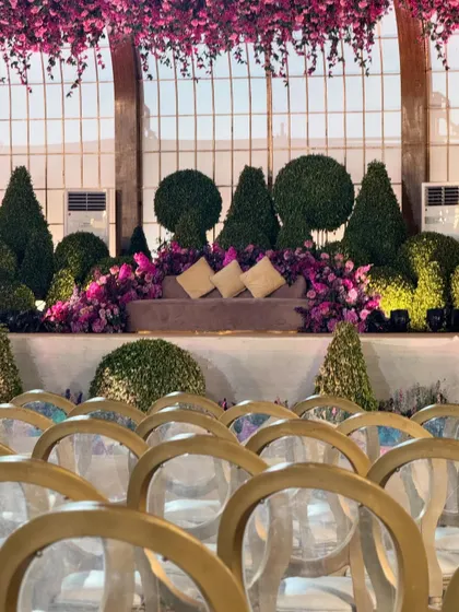 A gorgeous seating area set against a backdrop of a greenhouse-style window. The space is filled with sculpted topiaries and a profusion of purple and pink flowers.