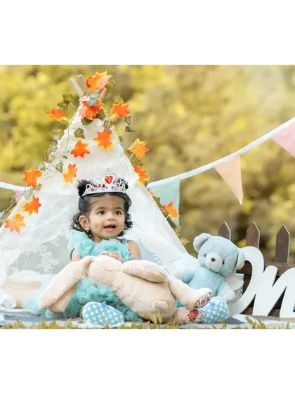 Princess Nitya looking adorable in her little tiara during her first birthday shoot. The setup is styled with soft toys and autumn leaves on the teepee to create a whimsical, fairytale feel.