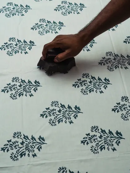 This image captures the heart of block printing: the moment of contact. An artisan's hand firmly presses a floral block onto fabric, transferring the design with practiced skill. It is in this simple, focused action that a plain cloth begins its transformation into a work of art.