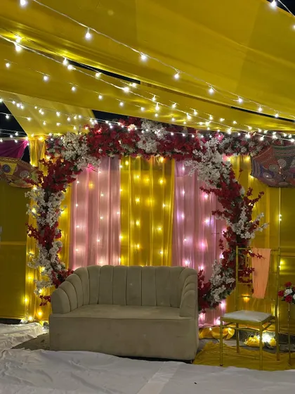 A bright and cheerful Haldi or Dholki backdrop on a terrace. The setup features a circular floral arch with red and white flowers against a backdrop of yellow and pink drapes lit by fairy lights.
