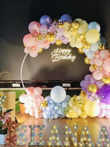 A dreamy pastel rainbow birthday setup with a multi-colored balloon arch, a 'Happy Birthday' neon sign, and marquee letters.
