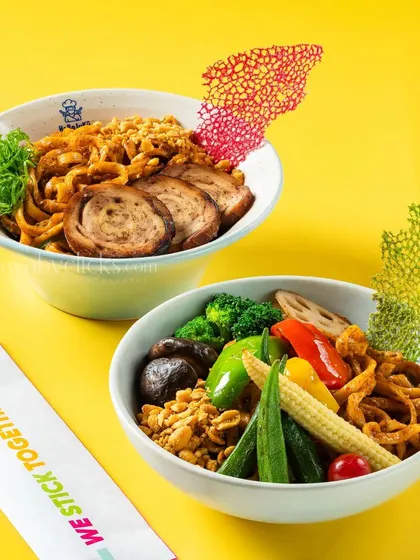 Two bowls of ramen, one with pork and the other with mixed vegetables, against a bright yellow background.