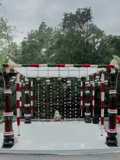 This mandap design is a beautiful blend of tradition and nature. The structure is decorated with red and white floral garlands wrapped around wooden pillars, with banana leaves adding an auspicious touch, perfect for a South Indian muhurtham.