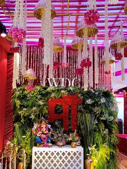 A beautiful setup for the ceremony, with an idol of Lord Ganesha placed on a jali-work console, set against a backdrop of lush greenery and a 'Maa' floral sign.