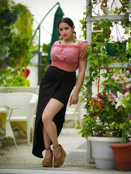 A model in a pink top and black slit skirt, posing in a lush, green cafe patio.