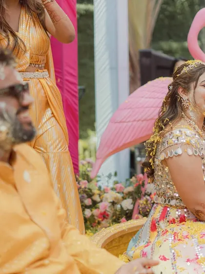 A candid shot of the bride laughing as she gets covered in haldi and flower petals. The playful flamingo prop in the background adds to the whimsical, pastel theme.