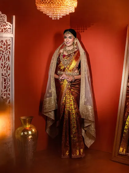 A full-view portrait showcasing a bride in a magnificent gold and maroon saree with a delicate veil. The studio setting with warm lights creates a royal ambiance.