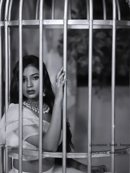 An artistic black and white photo of a model in a white gown, posing inside a large birdcage prop for a conceptual shoot.