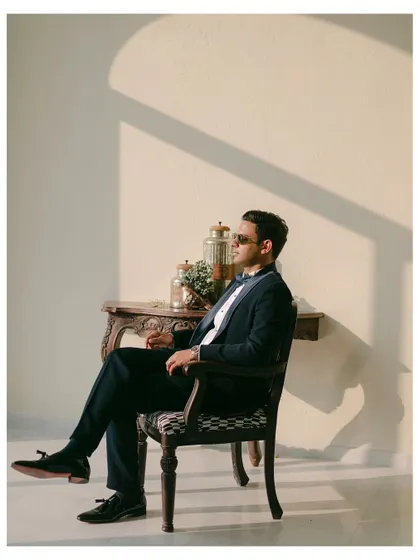 A groom sits in a vintage chair, bathed in the warm glow of the sun. This relaxed, stylish portrait captures a moment of quiet confidence before the ceremony.