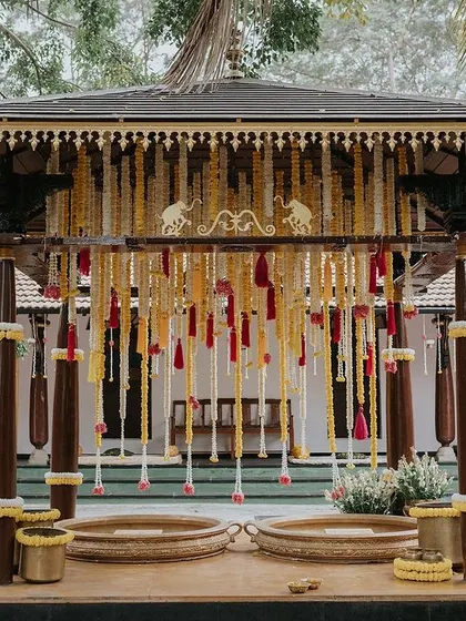 A frontal view of the Haldi mandap, decorated with yellow and white tuberose strings and colorful tassels. The two large brass urlis are ready for the ceremony's rituals.
