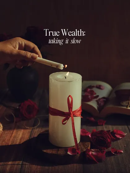 A moody, still-life photo with the theme "True Wealth: taking it slow." The scene of lighting a candle next to a book and roses evokes a sense of peace and mindfulness.