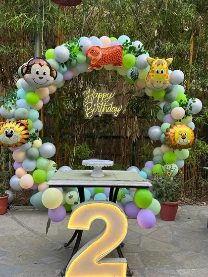 A fun and colorful jungle animal theme for a second birthday. This outdoor setup uses a circular balloon arch decorated with friendly animal foil balloons like a monkey, tiger, and giraffe, complete with a neon sign and a large marquee number '2'.
