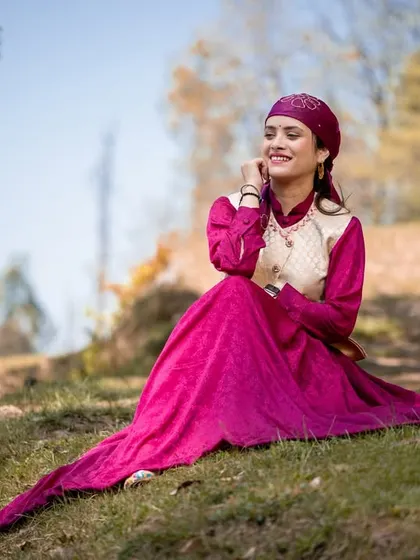 A graceful solo portrait of the woman in her flowing magenta Himachali dress, seated on a grassy slope.