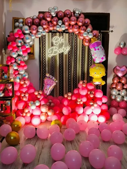 An 'Oh Baby' shower setup announcing "It's a Girl!". The room is filled with pink balloons, and the backdrop features a circular arch of pink and silver balloons with a neon sign.